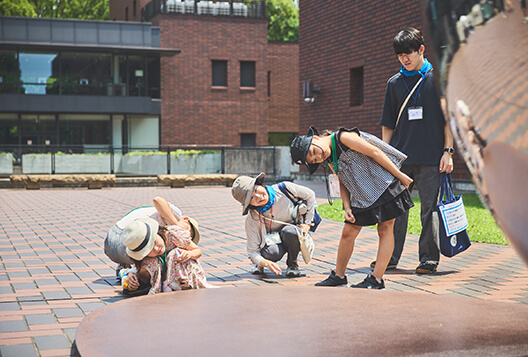 屋外彫刻を前に子どもたちととびラーが身をかがめて観察する様子