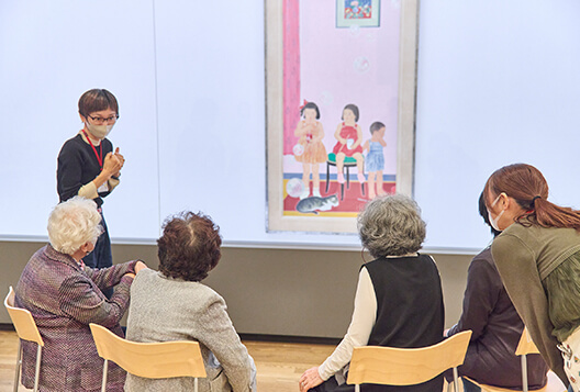 椅子に座る高齢者ととびラーが作品を見ながら穏やかに対話する様子