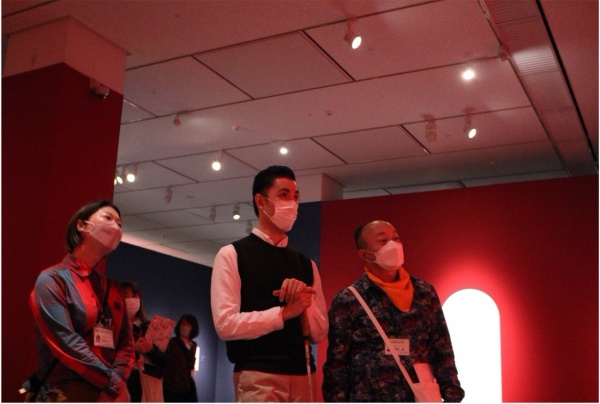 美術館の展示室の中で３人の人が作品を鑑賞している写真。展示室の壁は赤い。真ん中の人は白杖を持っている。