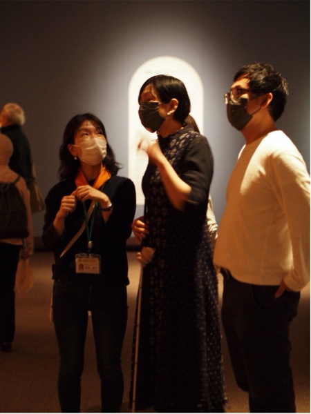 展示室で３人の人が作品を鑑賞している写真。真ん中の人は白杖を持っている。